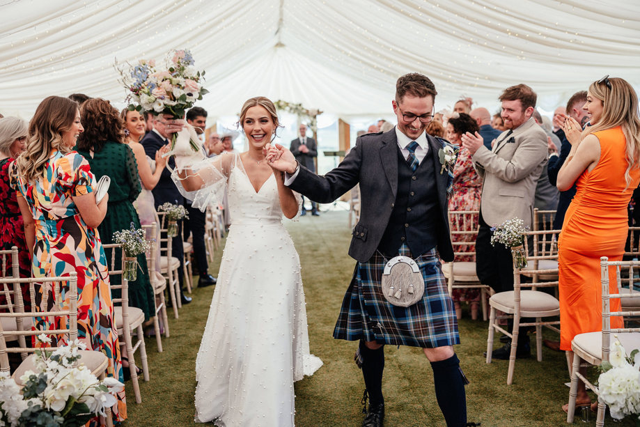 Bride and groom holding hands and smiling as they walk back down the aisle together as guests clap